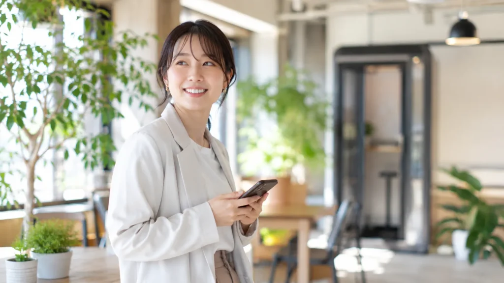 自然光が差し込む明るいコワーキングスペースで、スマホを手にしながら快適そうに過ごす日本人女性。記事全体の「快適なワーク環境」を表現しています。