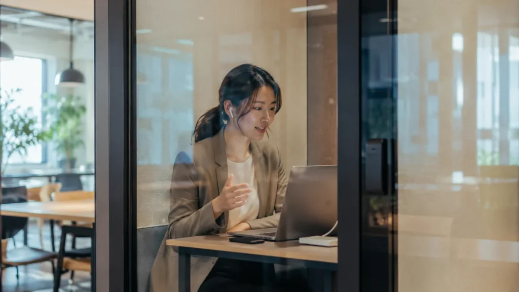 ガラス張りの防音ブース内で快適にWeb会議を行う日本人ワーカーと静かに整えられたコワーキングフロア